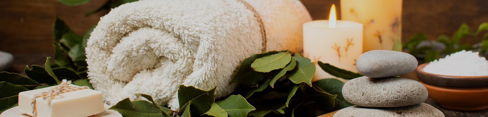 Spa essentials with a rolled towel, stacked stones, candles, a soap bar, and green leaves creating a serene ambiance.